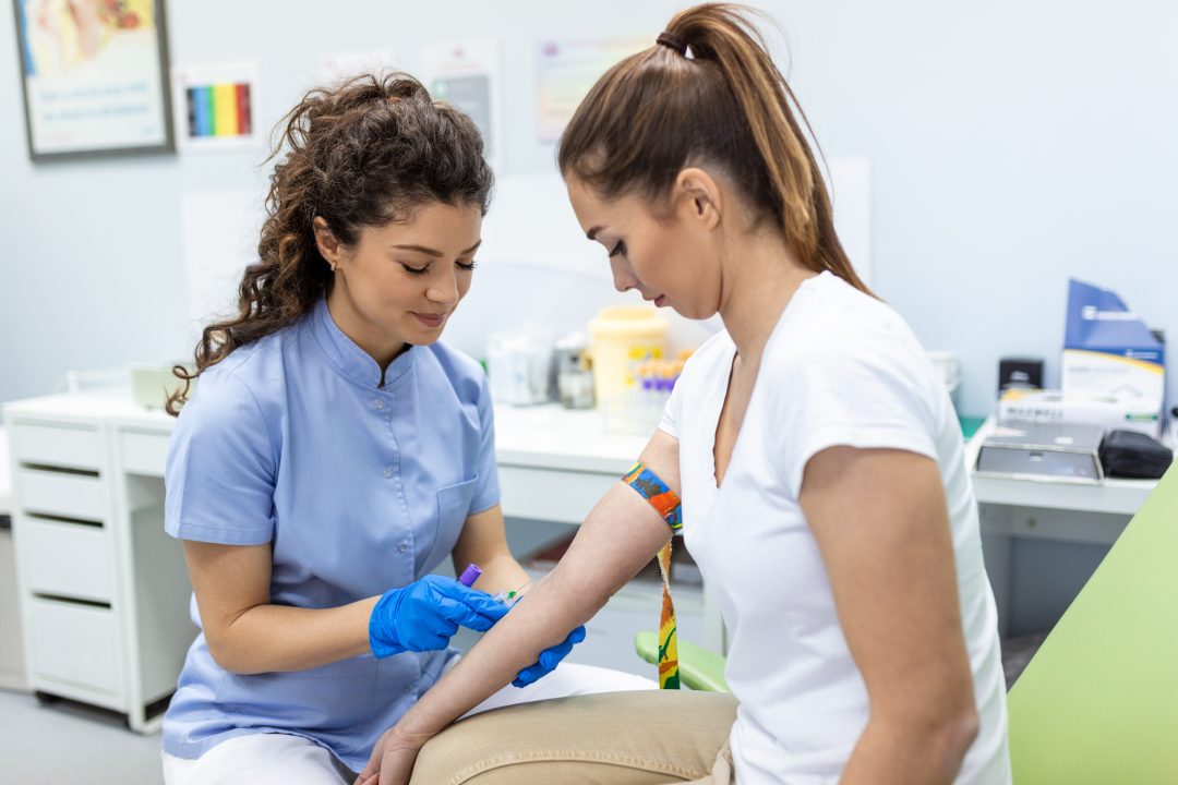 Preparación de un análisis de sangre con una mujer joven y guapa por una doctora con uniforme médico sobre una mesa en una habitación blanca y luminosa. La enfermera perfora la vena del brazo de la paciente con un tubo blanco con aguja.