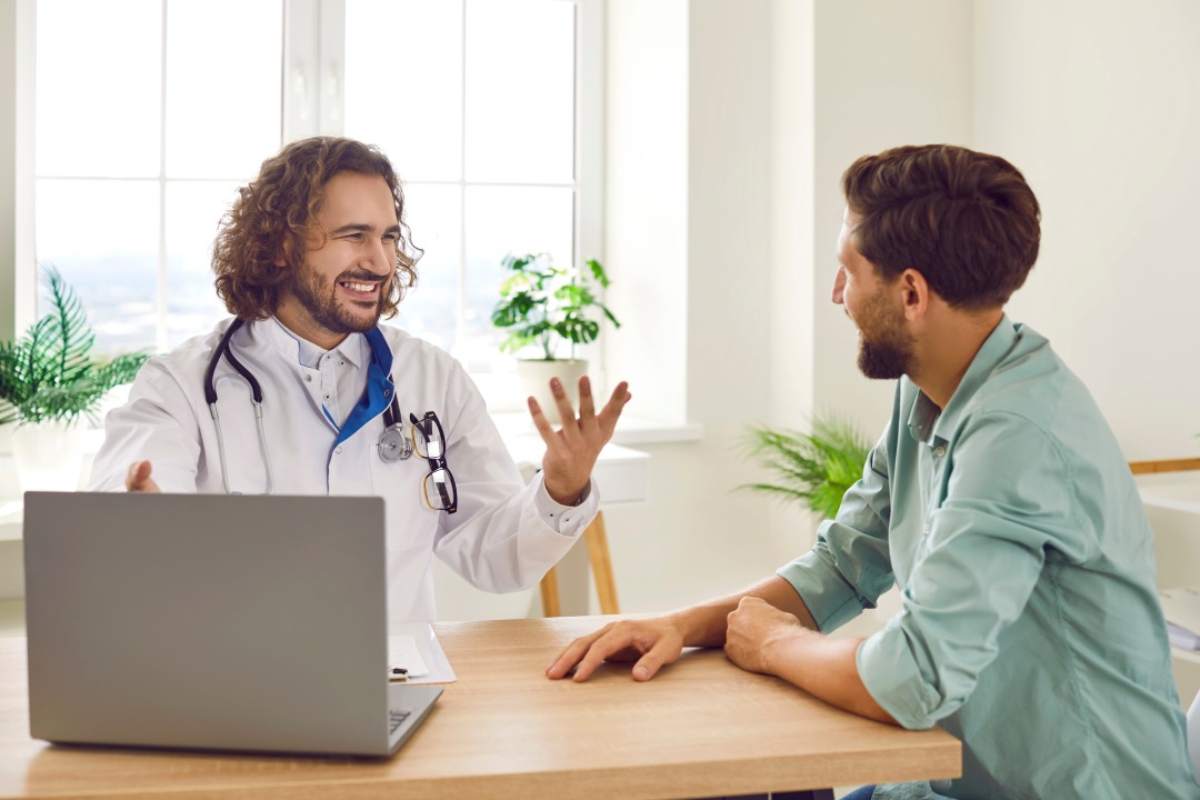 Retrato de un paciente joven sonriente y feliz en la consulta escuchando las recomendaciones de un amable médico sentado en el escritorio de una clínica durante un examen médico. Concepto de atención sanitaria.