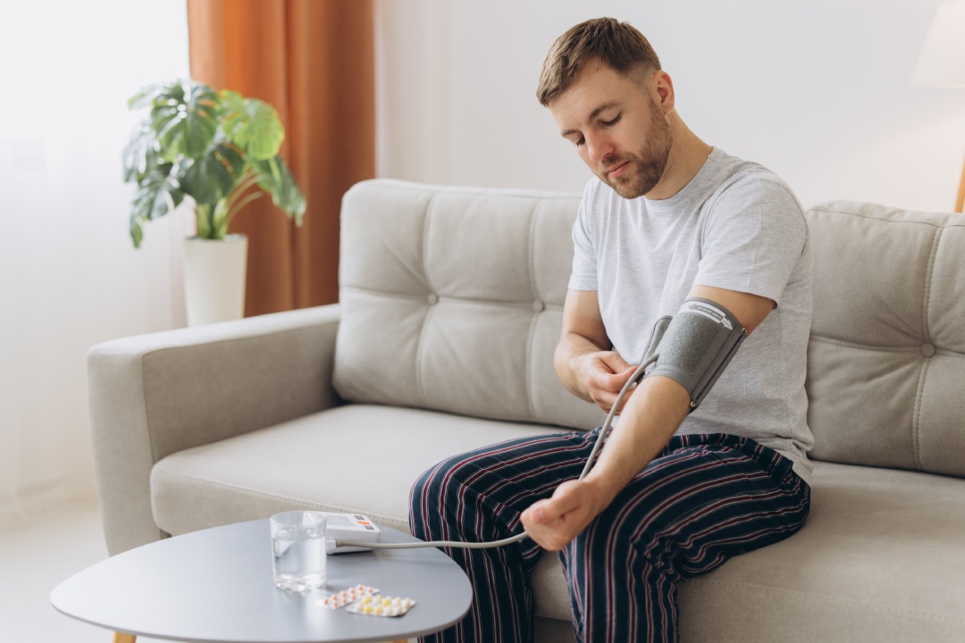 Young caucasian man sitting on couch at home, checking blood pressure. Bearded guy in pajamas using modern tonometer, experiencing hypertension caused by stress and headache, copy space