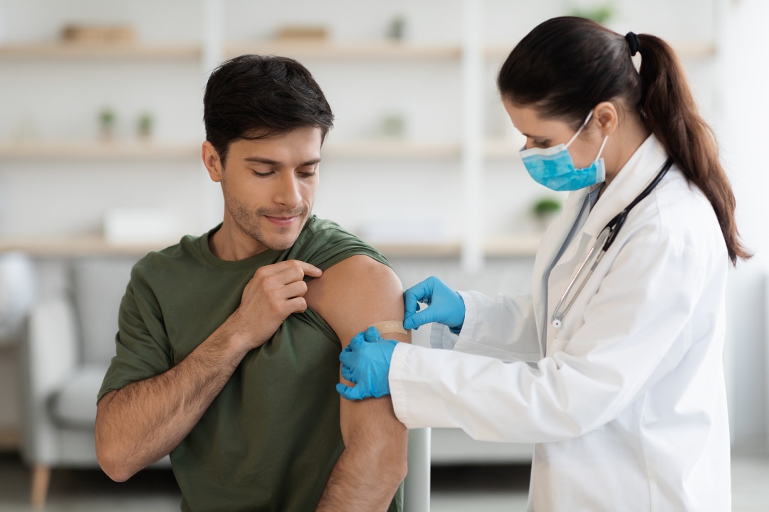 Millennial man in camouflage uniform soldier getting vaccinated against coronavirus at clinic while pandemic, female doctor in protective workwear making shot in shoulder for military guy